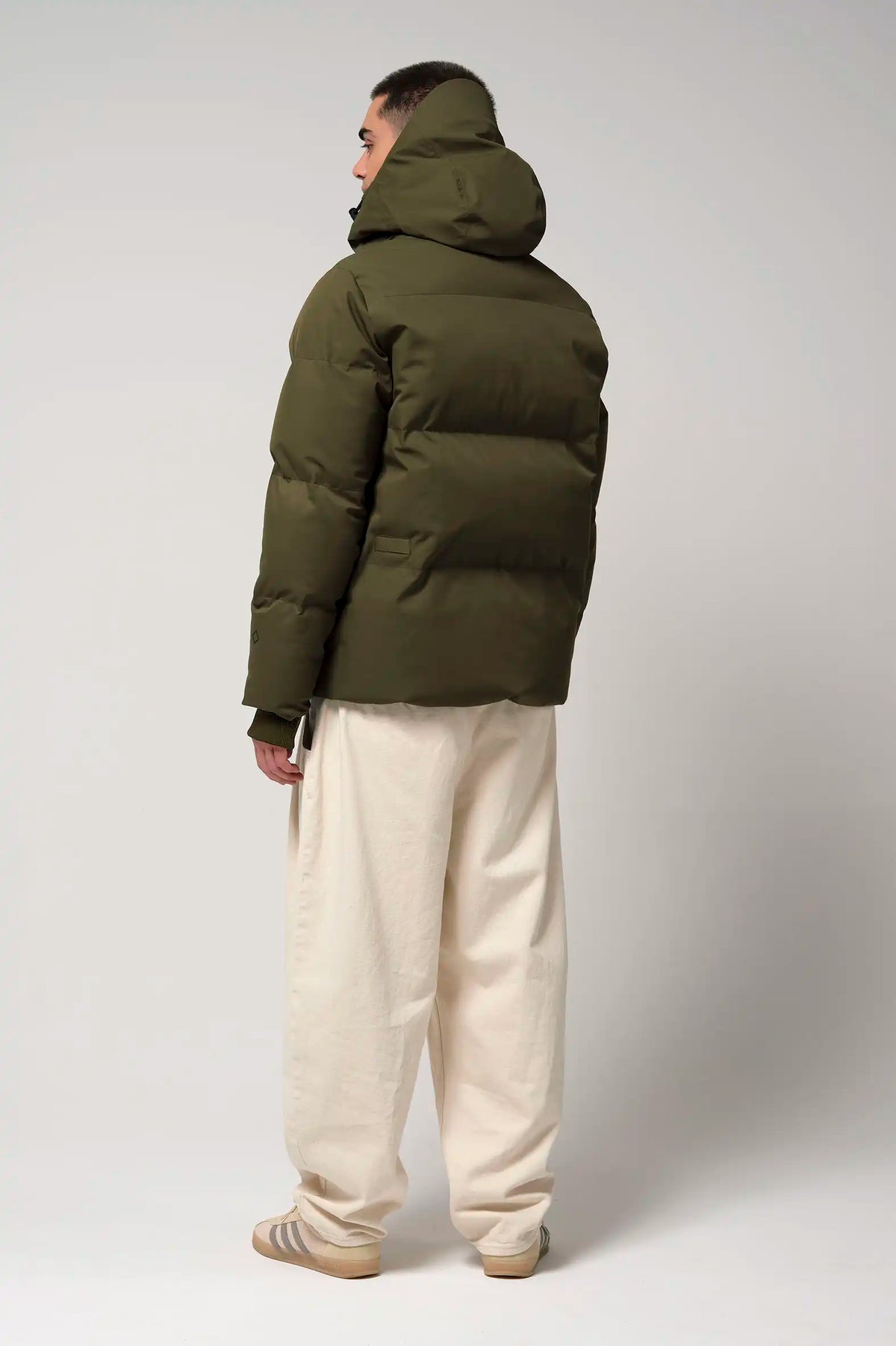 Bayu Dark Olive Green Coat shown from the back, featuring a padded design and hood, worn with light pants.