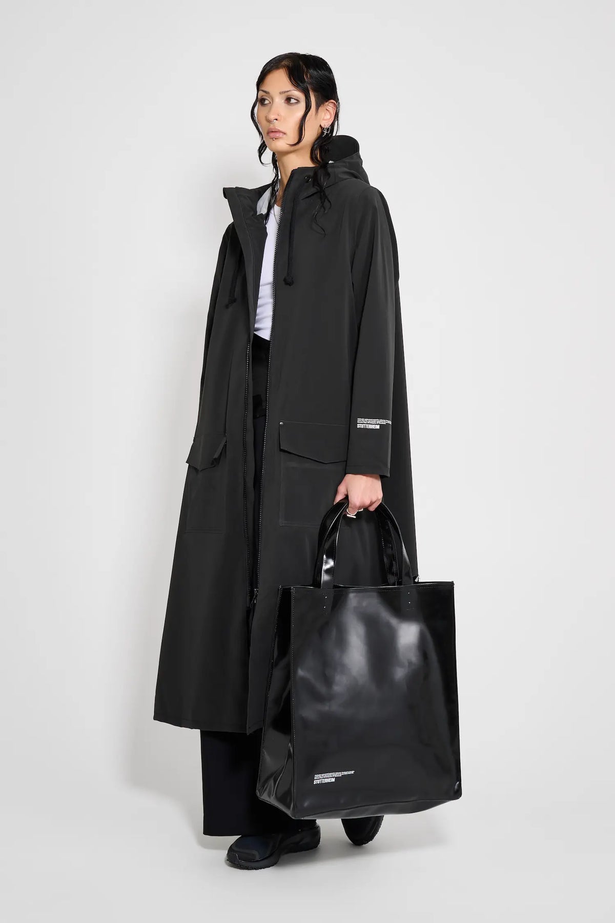 Model wearing Mosebacke Long Patch Black Raincoat with adjustable hood and black tote bag.