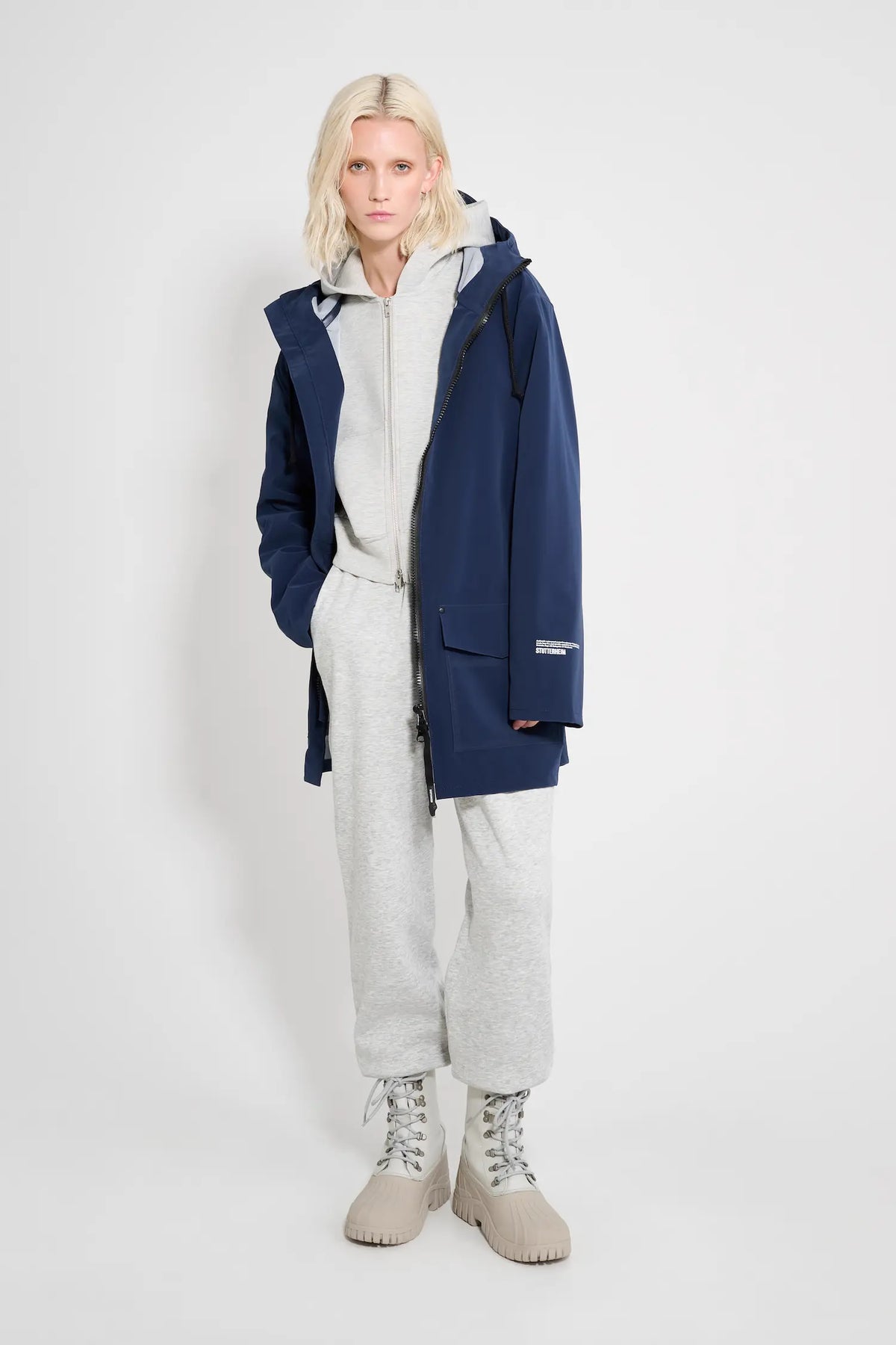Model wearing the Stockholm Patch Navy Raincoat, a completely waterproof oversized rain jacket, styled with gray pants and boots.