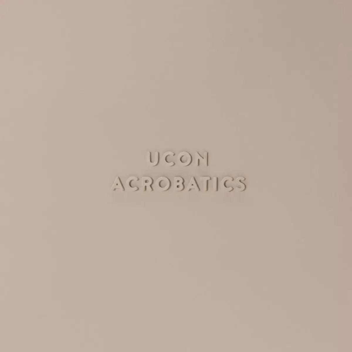 Logo of UCON ACROBATICS on a soft beige background, representing stylish and functional bag designs.