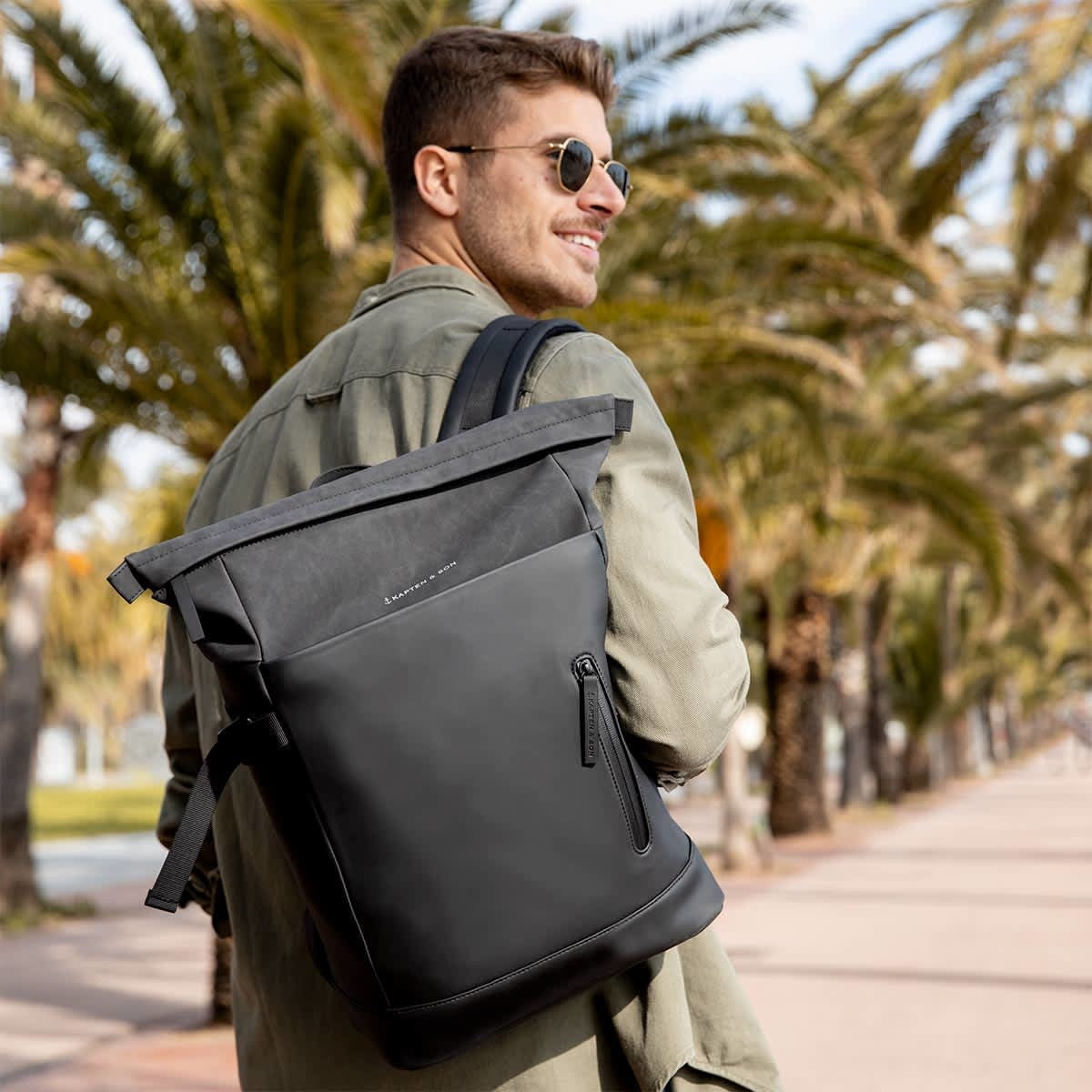 Man wearing a Helsinki Sandstone Backpack while walking on a palm-lined path, showcasing its stylish design.