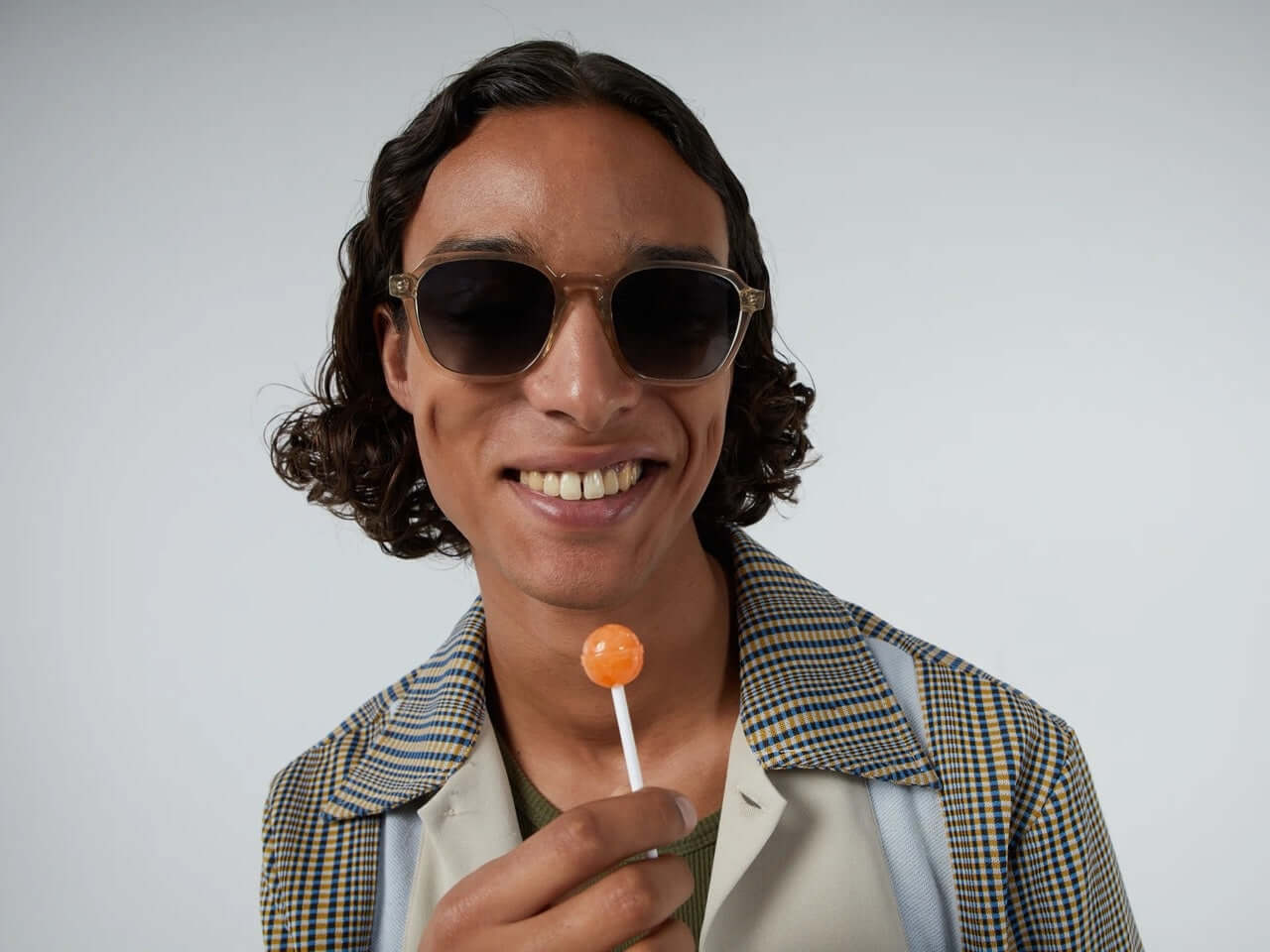Matty Blue Sands Sunglasses model smiling with lollipop, wearing stylish sunglasses against a gray background.