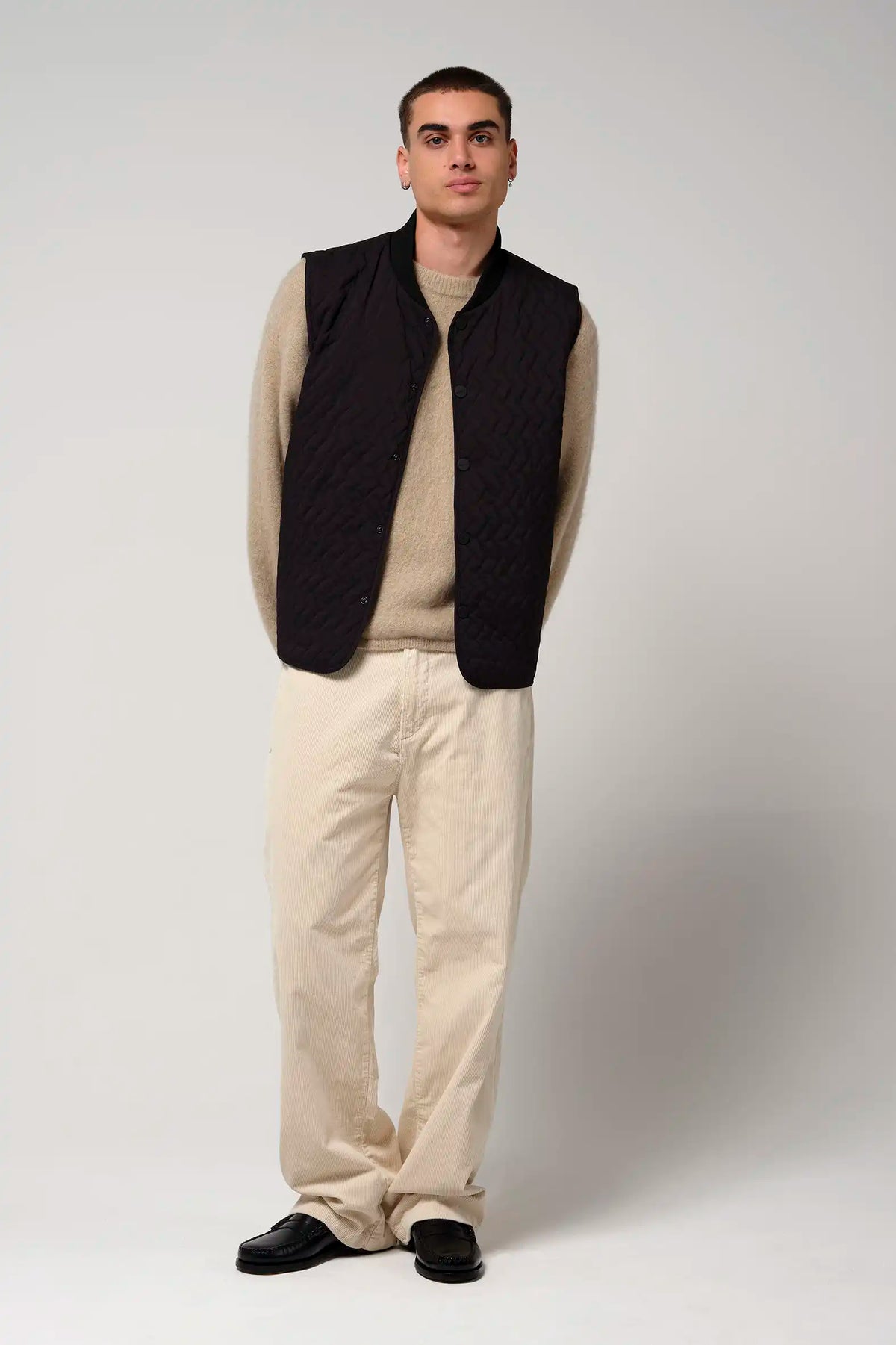 Model wearing a Claud Deep Black Coat vest, paired with a beige sweater and white pants, against a neutral background.