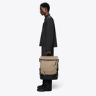 Side view of a person carrying a Trail Mountaineer Rolltop Beige Backpack, showcasing its stylish design and functionality.