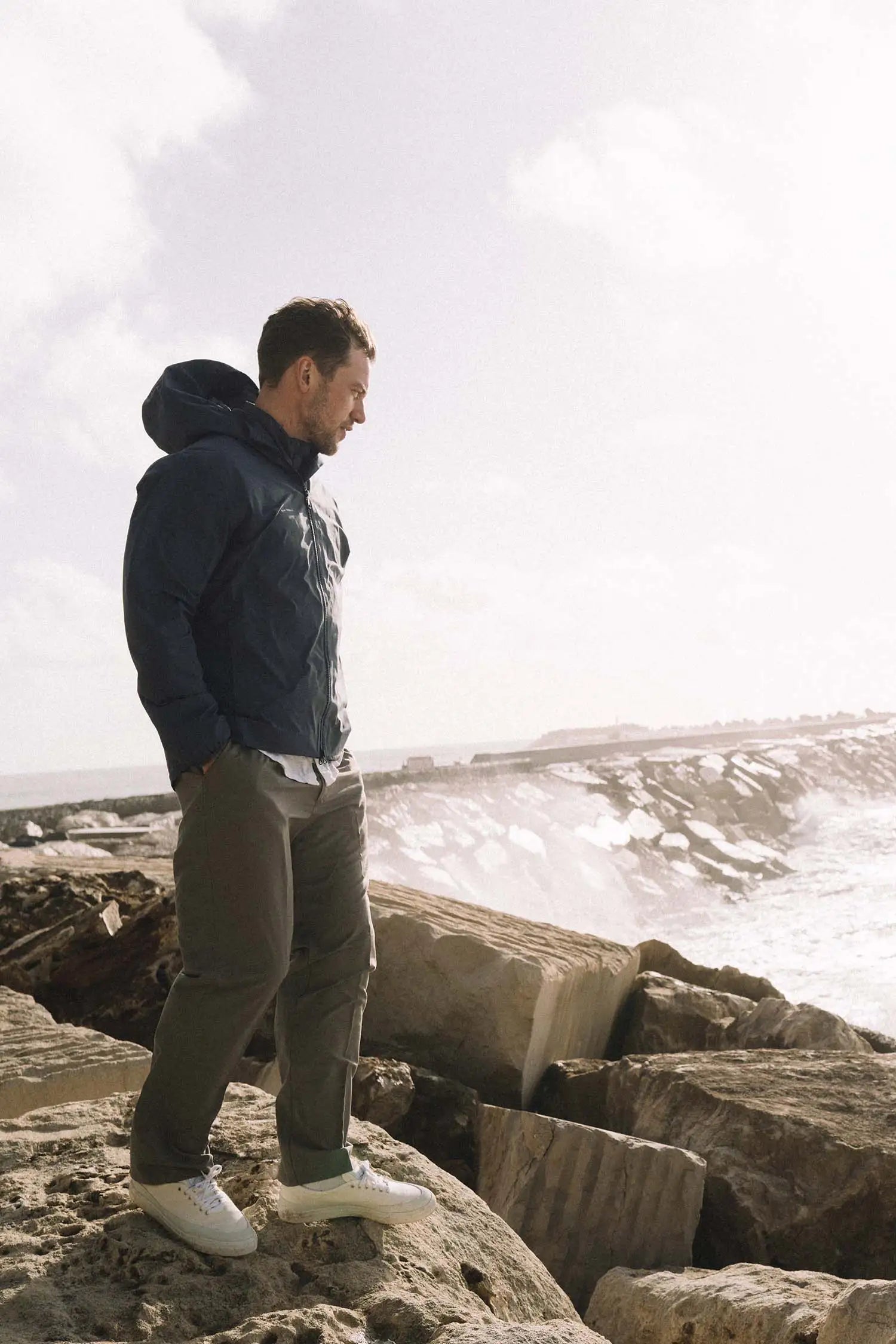 Man wearing Hood II Midnight Jacket standing on rocks by the ocean with waves in the background.