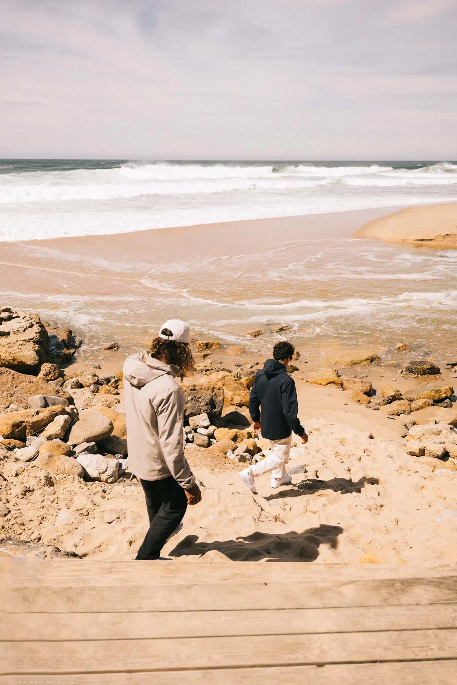 Two individuals walking along a beach near waves, wearing casual attire and enjoying the coastal scenery.