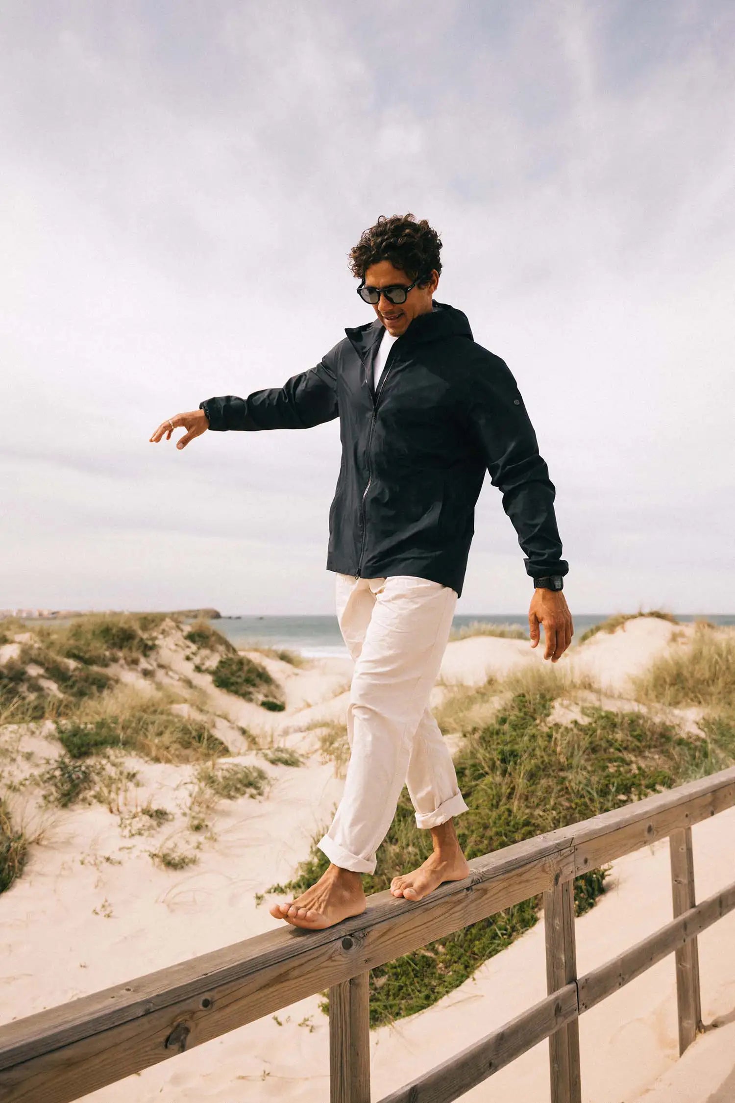 Man wearing the Hood II Midnight Jacket walking on a wooden railing at the beach on a cloudy day.