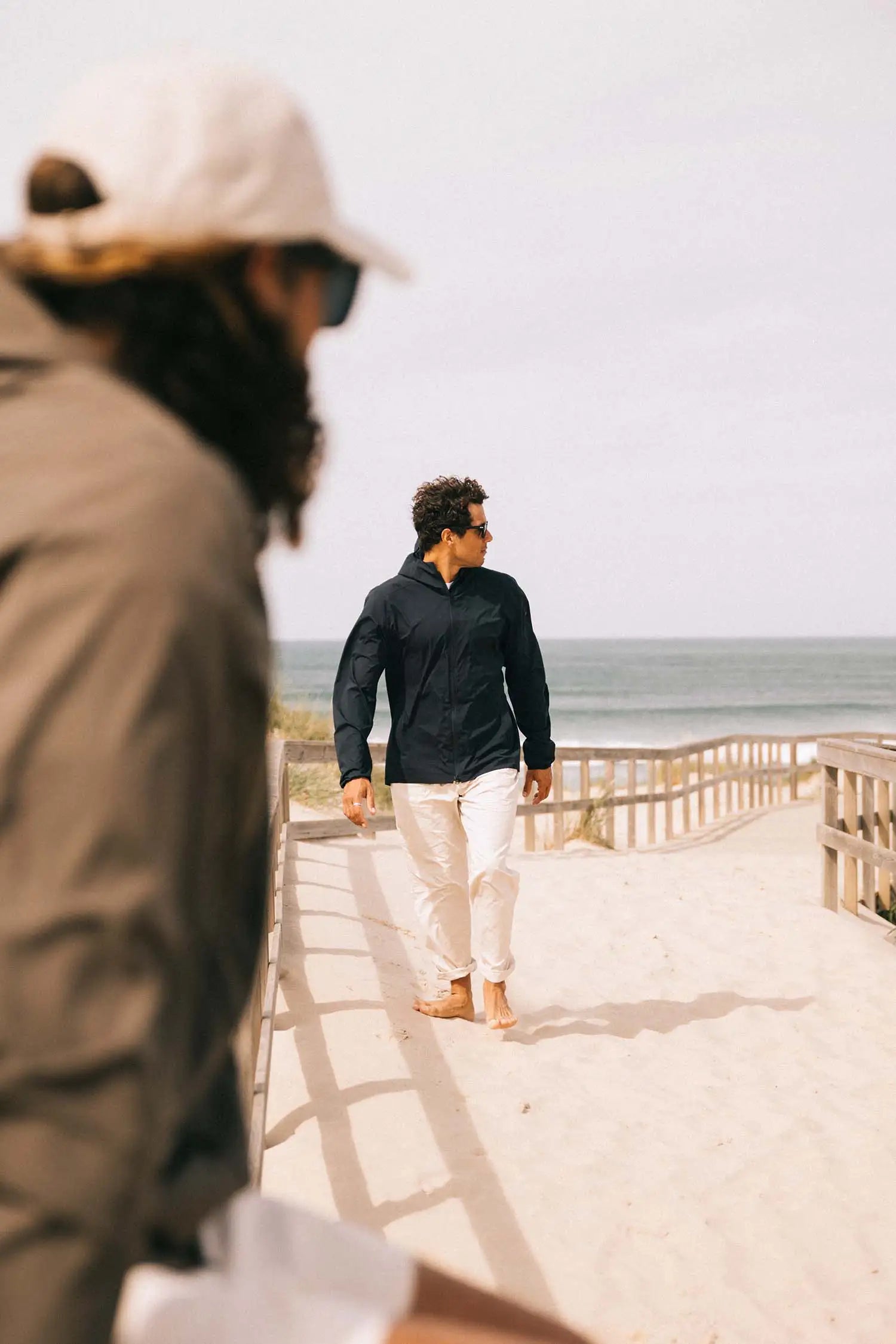 Man in Hood II Midnight Jacket walking on beach path with sea in background, enjoying lightweight comfort.