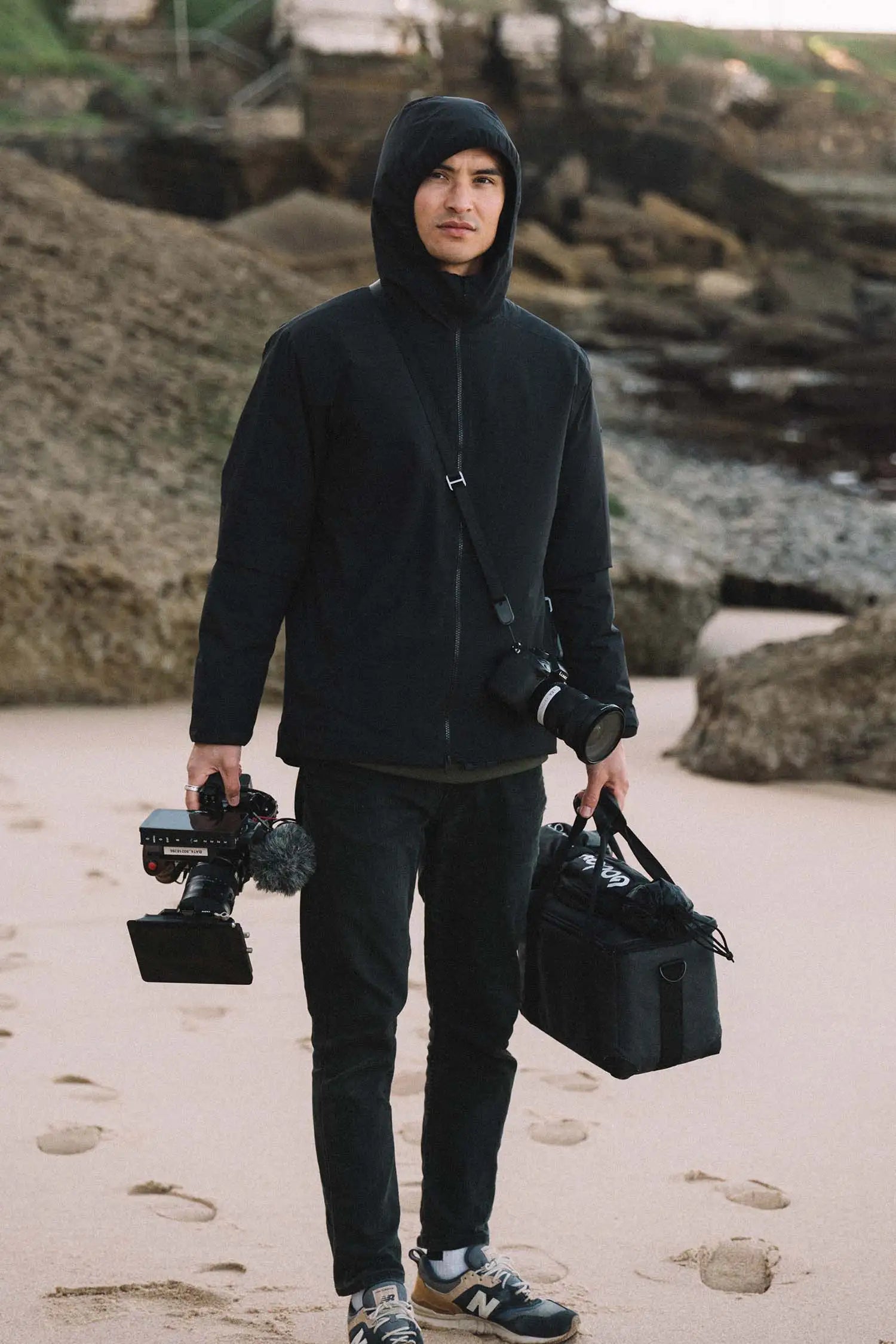 Person wearing a Hood II Onyx Jacket on the beach, holding camera gear and a bag.