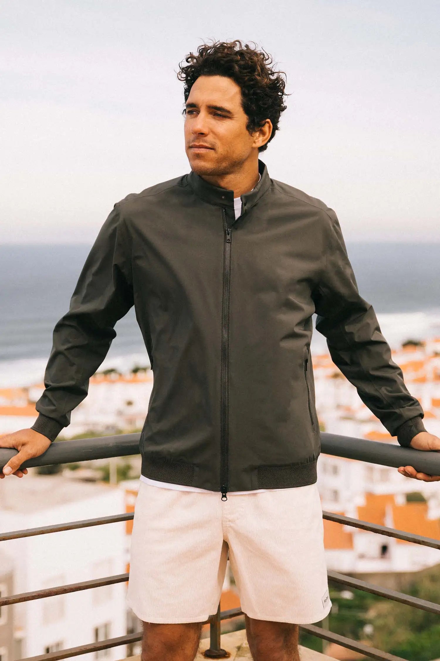Model wearing a Plain II Dark Olive Jacket on a balcony with a coastal view.