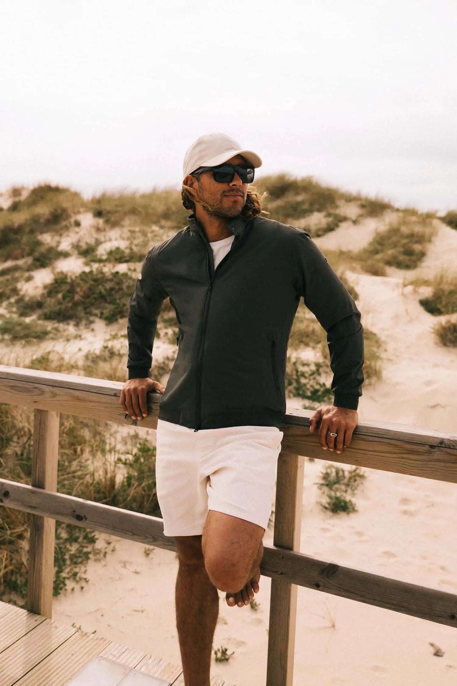 Man wearing a Plain II Dark Olive Jacket, enjoying an outdoor setting near sandy dunes.