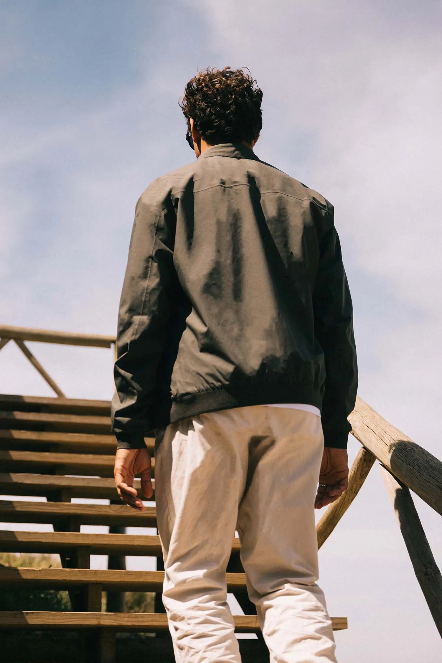 Man wearing a Plain II Dark Olive Jacket walking up wooden stairs under a clear sky.