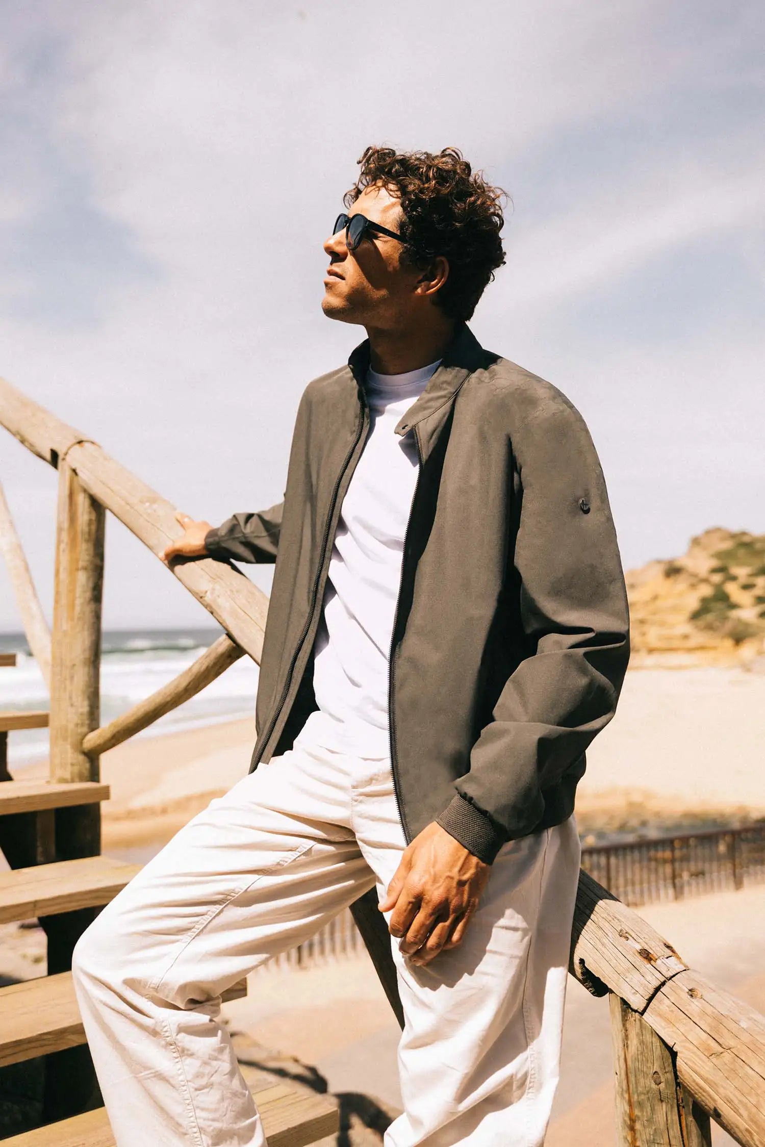 Man wearing a stylish Plain II Dark Olive Jacket on a sunny beach, showcasing its versatile summer design.