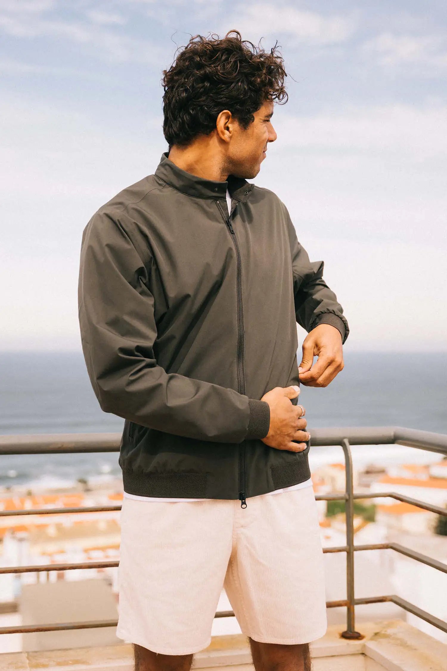 Man wearing Plain II Dark Olive Jacket against a coastal background, showcasing a modern summer style.