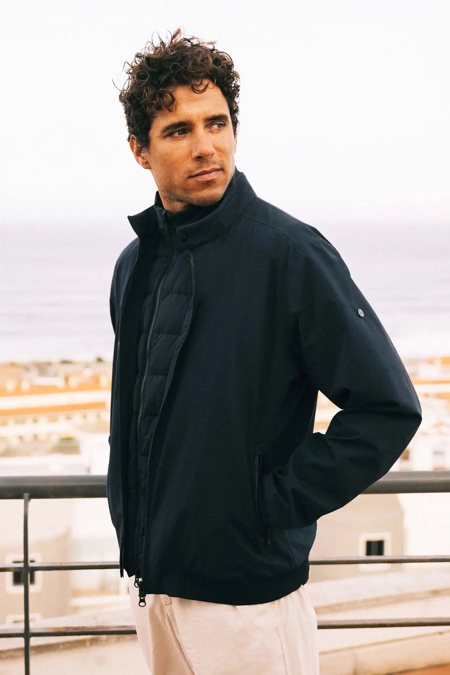 Model wearing Plain II Midnight Jacket with modern city look against scenic ocean background.