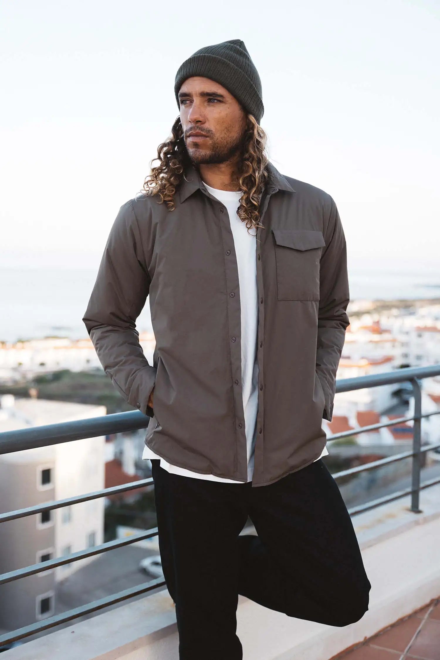 Model wearing Trip II Major Brown Overshirt with beanie, standing on balcony overlooking the seaside.