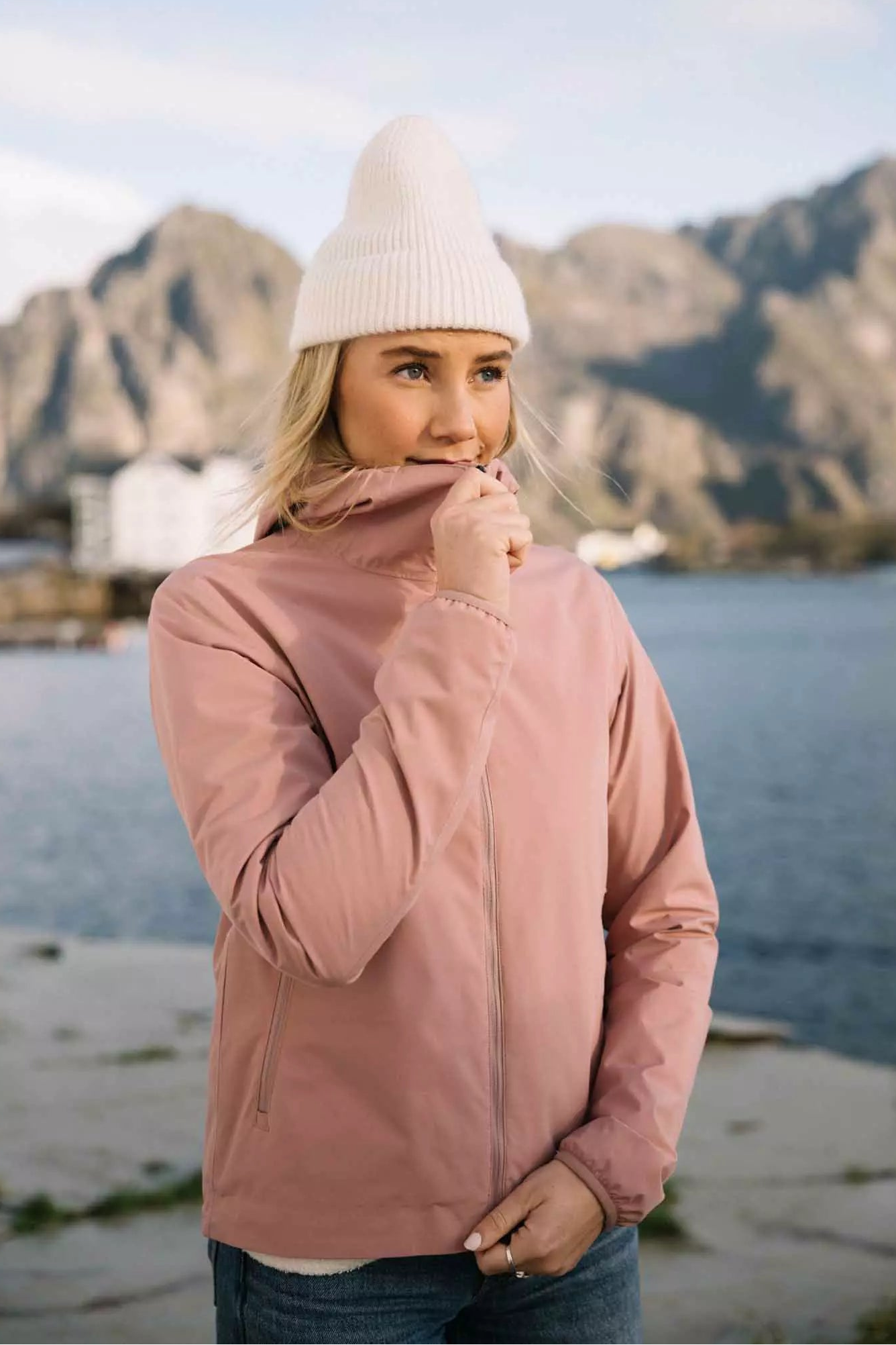 Veil II Flush Pink Jacket model wearing a pink jacket with a white beanie by the water in a scenic outdoor setting.