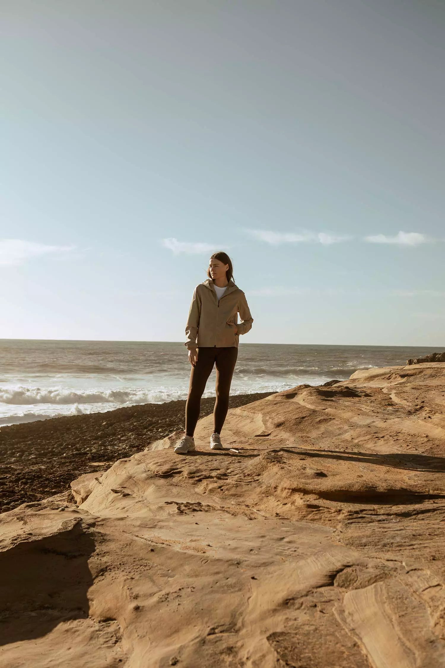 Woman wearing Veil II Khaki Jacket standing on a rocky beach by the ocean.