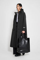 Model wearing Mosebacke Long Patch Black Raincoat with adjustable hood and black tote bag.