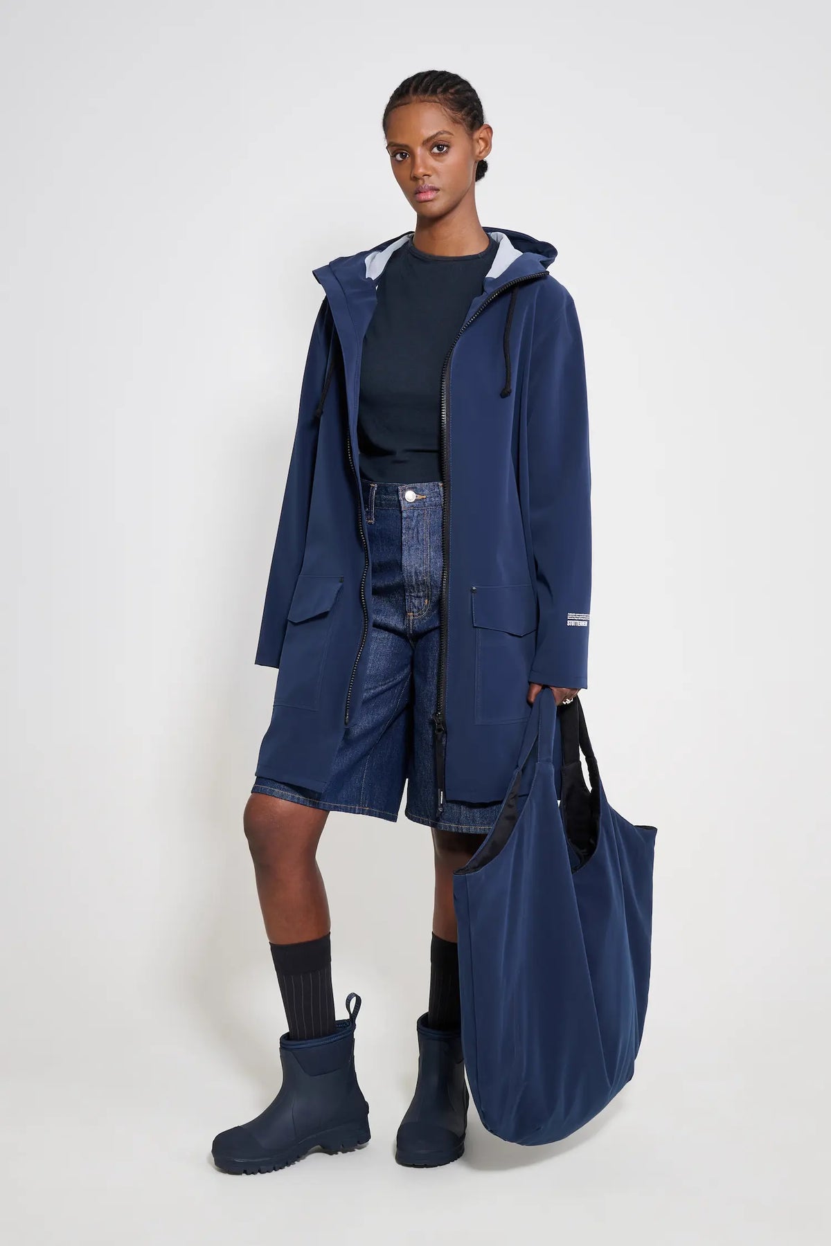 Model wearing Mosebacke Patch Navy Raincoat with adjustable hood, paired with shorts and boots.