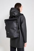 Model wearing Rain Rolltop 25L Black Backpack, showcasing its waterproof design and rolltop opening.