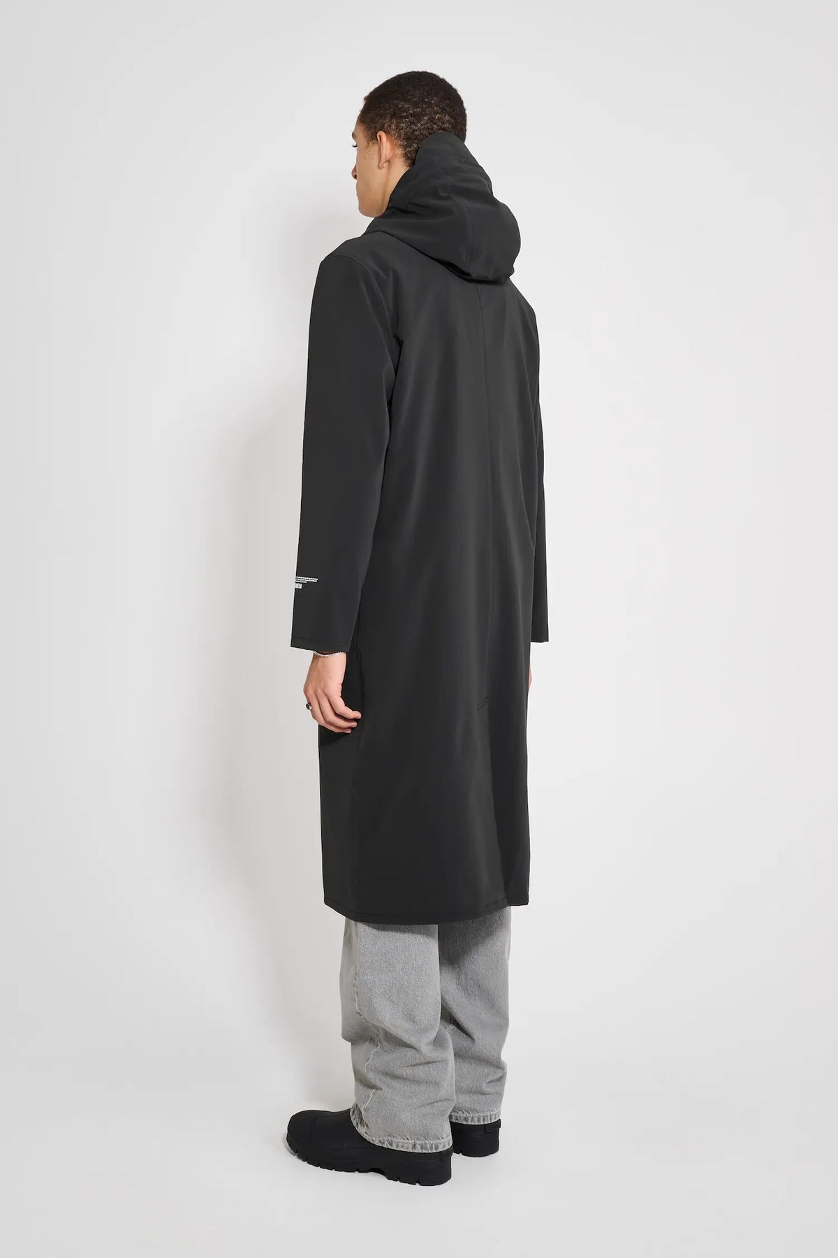 Back view of a model wearing the Stockholm Long Patch Black Raincoat with a drawstring hood and oversized fit.