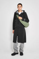 Model wearing Stockholm Long Patch Black Raincoat with an adjustable drawstring hood and green bag.