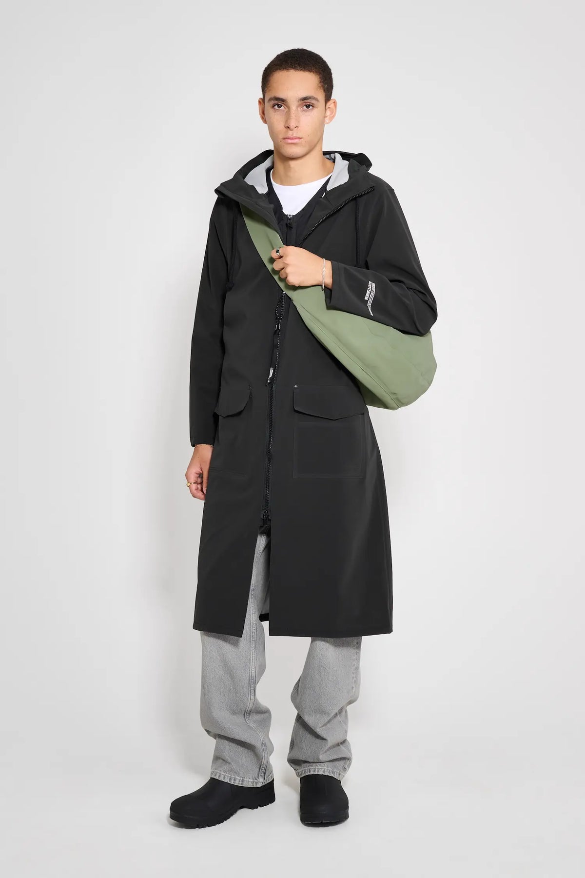 Model wearing Stockholm Long Patch Black Raincoat with an adjustable drawstring hood and green bag.