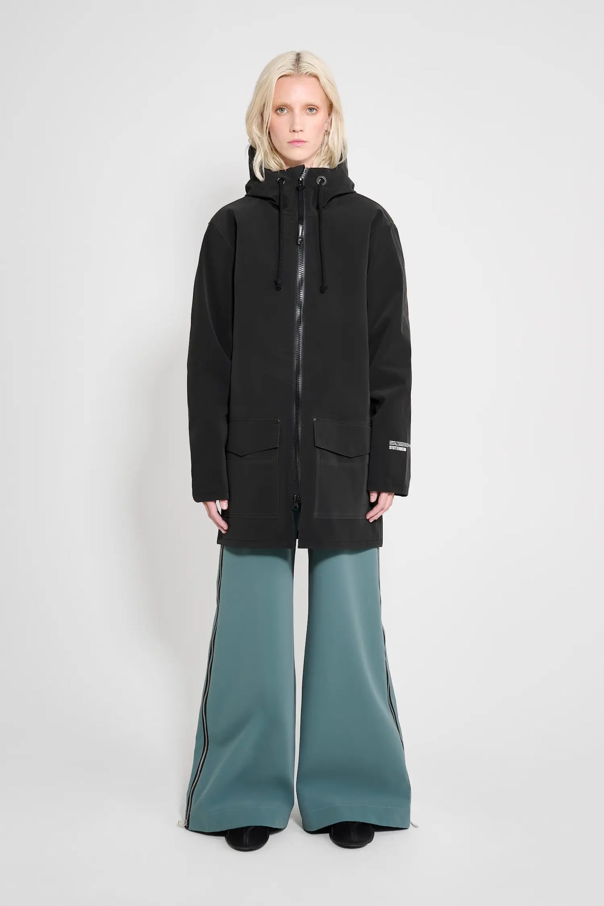 Model wearing the Stockholm Patch Black Raincoat in black, paired with stylish wide-leg pants.